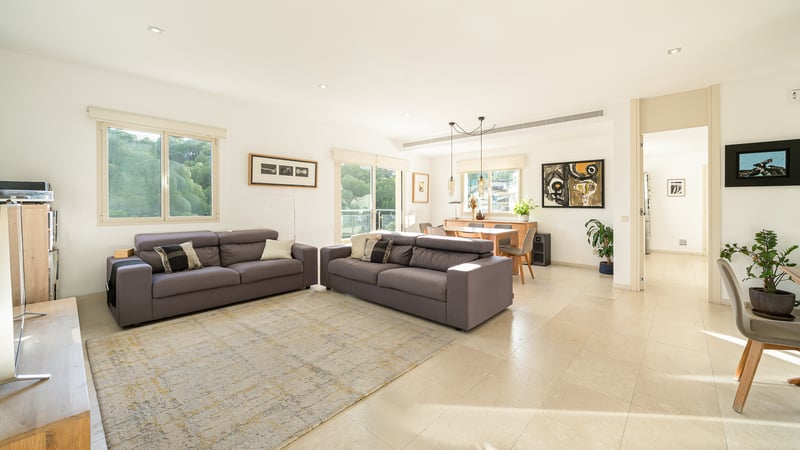 Grand salon lumineux avec deux canapés gris, carrelage clair et coin repas en arrière-plan avec de grandes fenêtres.
