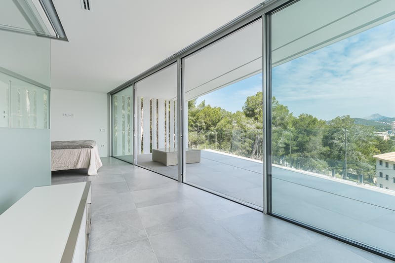 Chambre minimaliste avec de grandes baies vitrées coulissantes ouvrant sur une terrasse surplombant la forêt.