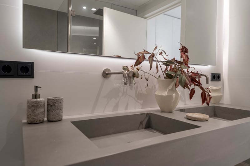 Double vasque grise intégrée avec robinetterie murale et miroir rétroéclairé dans une salle de bain moderne.