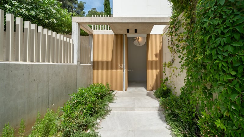 Entrée de villa moderne avec lattes de bois verticales, murs en béton lisse et végétation luxuriante.