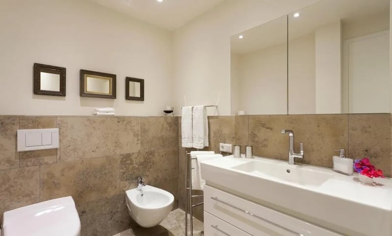 Salle de bain moderne avec carrelage en pierre beige, meuble vasque blanc, bidet et grand miroir mural.