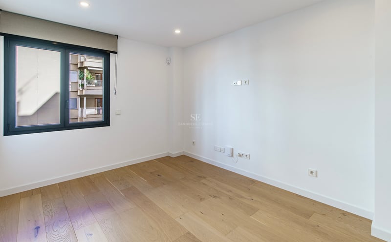 Habitación vacía y luminosa con suelos de madera clara, paredes blancas y un gran ventanal con persiana.