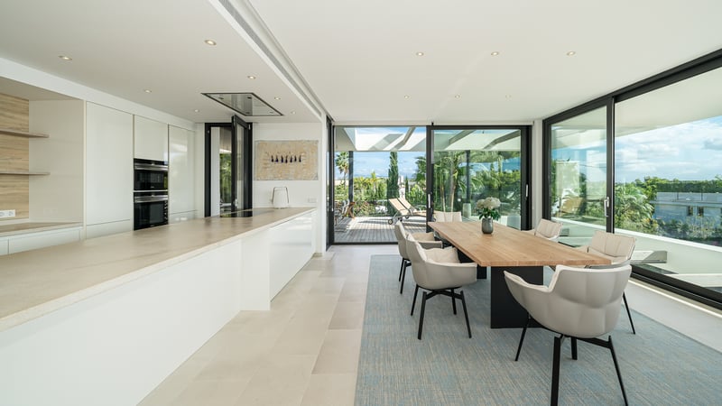 Salle à manger moderne avec table en bois, chaises grises et cuisine blanche, ouverte sur une terrasse par des baies vitrées.