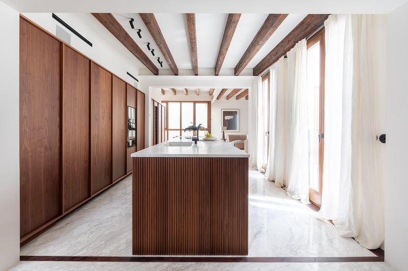 Cocina de lujo con isla de madera alistonada, encimeras de mármol y vigas de madera oscura bajo luz natural.