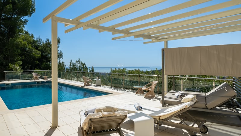 Terrasse ensoleillée avec piscine, pergola blanche, transats et garde-corps en verre surplombant la mer.