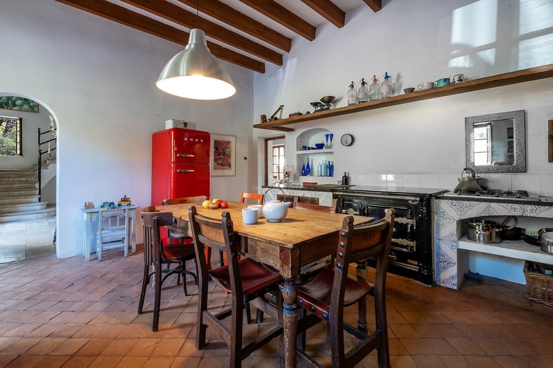 Cuisine traditionnelle avec sols en terre cuite, poutres en bois, table rustique et réfrigérateur rouge vif.