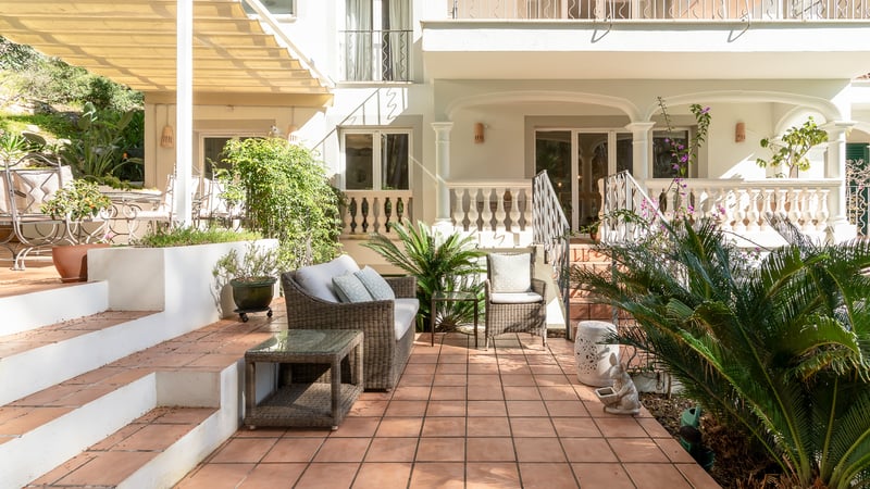 Terrasse ensoleillée en terre cuite avec mobilier en osier et plantes devant une façade de villa blanche.