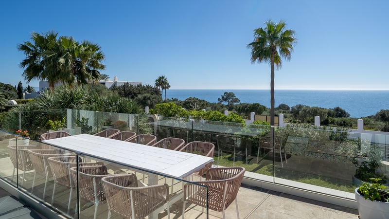 Grande terrasse avec table blanche, chaises design et garde-corps en verre surplombant la mer Méditerranée et des palmiers.