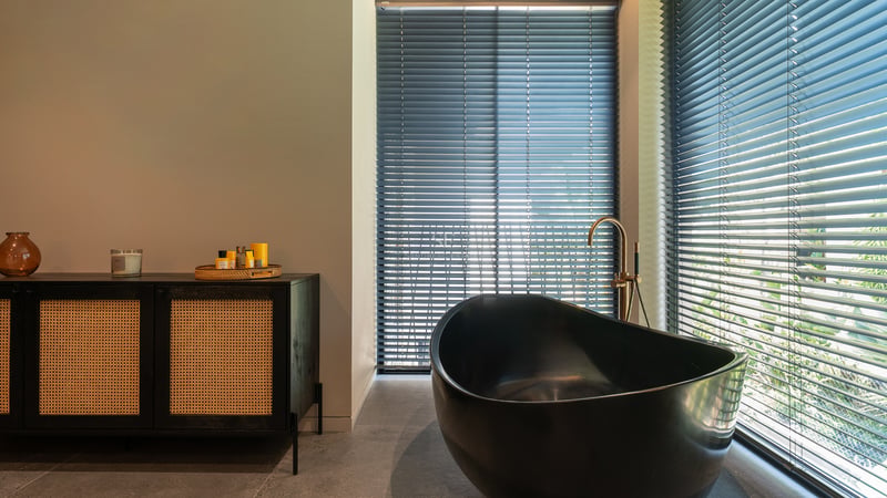 Salle de bain de luxe avec baignoire ovale noire, robinetterie dorée et buffet en rotin près de fenêtres à stores.