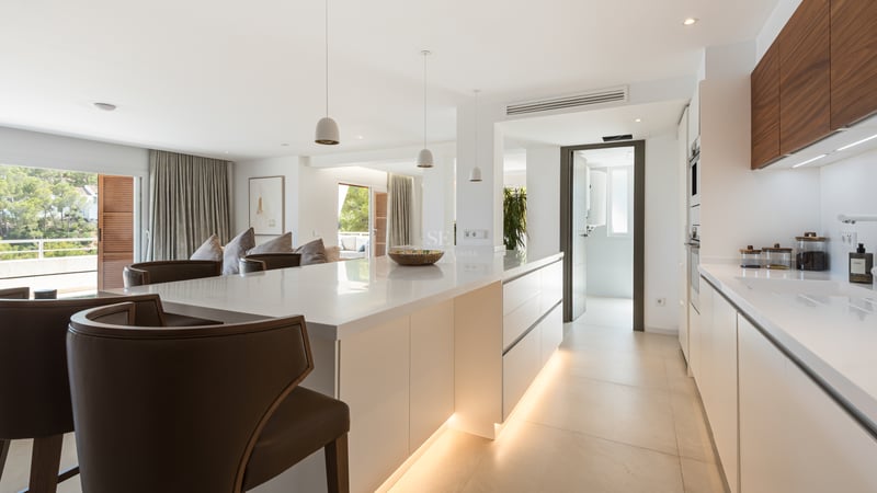 Cuisine moderne lumineuse avec grand îlot blanc, tabourets en cuir et menuiserie en noyer intégrée.