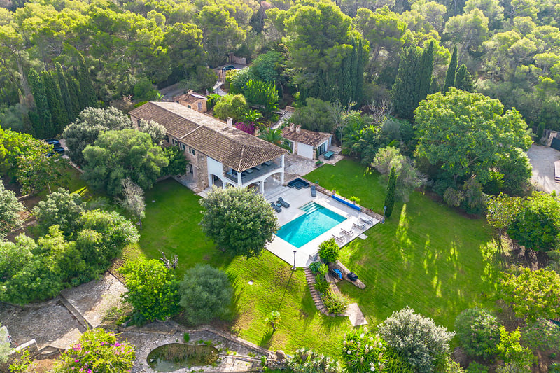 Vue aérienne d'une grande villa en pierre entourée d'une forêt, avec piscine turquoise et pelouses vertes.