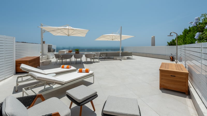 Grande terrasse blanche avec bains de soleil, coin repas, douche extérieure et garde-corps en verre face à la mer.