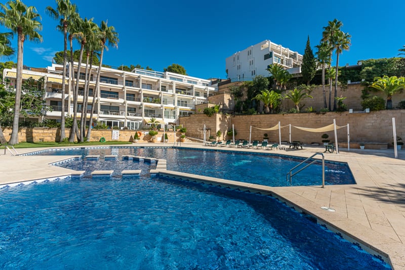 Grande piscine bleue entourée d'une terrasse en pierre, de palmiers et d'un immeuble blanc sous un ciel dégagé.