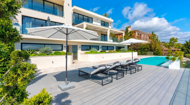 Extérieur d'une villa moderne avec piscine turquoise, chaises longues grises et parasols blancs dans un cadre paysager.
