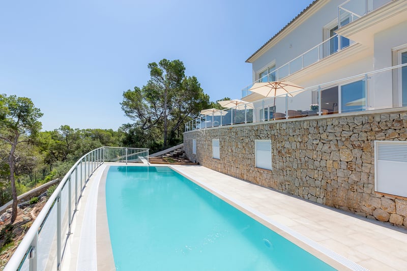 Une longue piscine turquoise bordant une villa moderne blanche avec des murs en pierre et des garde-corps en verre.