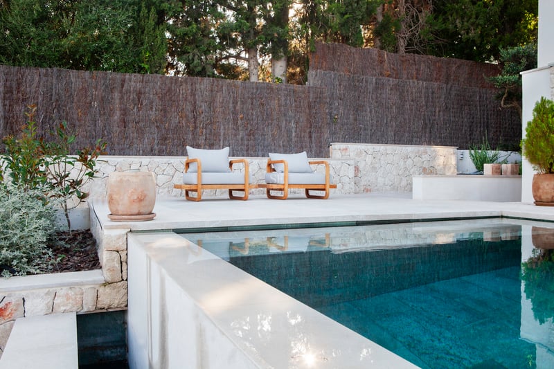 Piscine à débordement aux eaux turquoise jouxtant une terrasse en pierre calcaire avec deux fauteuils en bois.