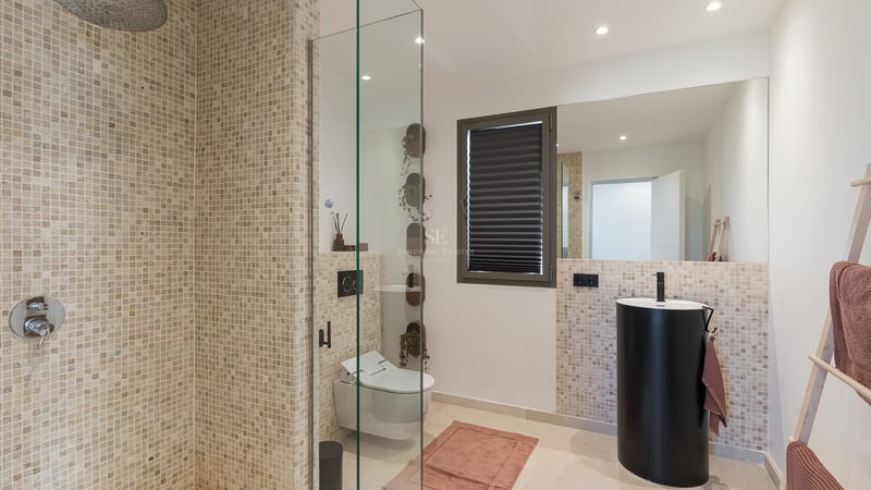 Salle de bain moderne avec mosaïque, paroi en verre, vasque colonne noire mate et toilettes suspendues.