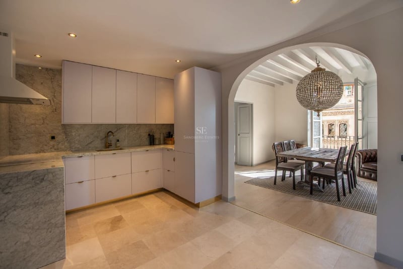 Cocina moderna blanca con encimeras de mármol que conecta mediante un arco con un comedor de madera y lámpara de cristal.