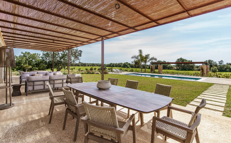 Gran mesa de comedor de madera bajo pérgola de cañizo en terraza de piedra frente a una piscina y jardín.