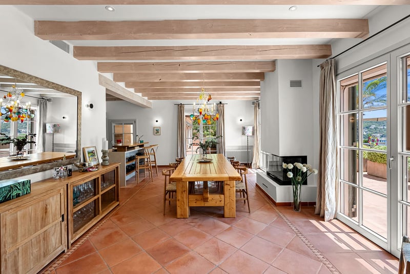 Grande salle à manger avec table en bois, sol en terre cuite et poutres au plafond menant à la terrasse.