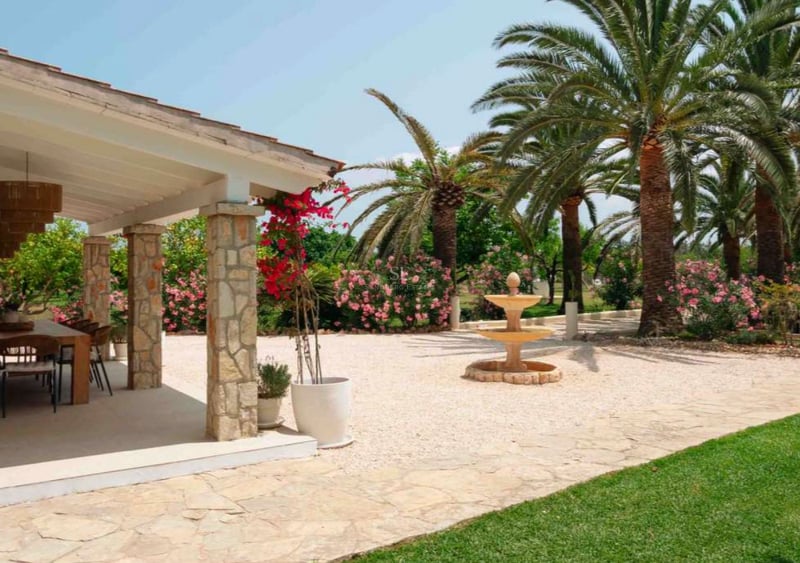Jardin méditerranéen avec terrasse en pierre, fontaine à trois niveaux et palmiers sous un ciel bleu.
