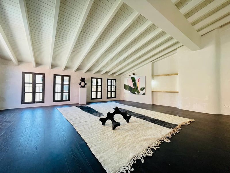 Grande pièce avec parquet foncé, plafonds à poutres blanches et un grand tapis texturé avec sculpture moderne.
