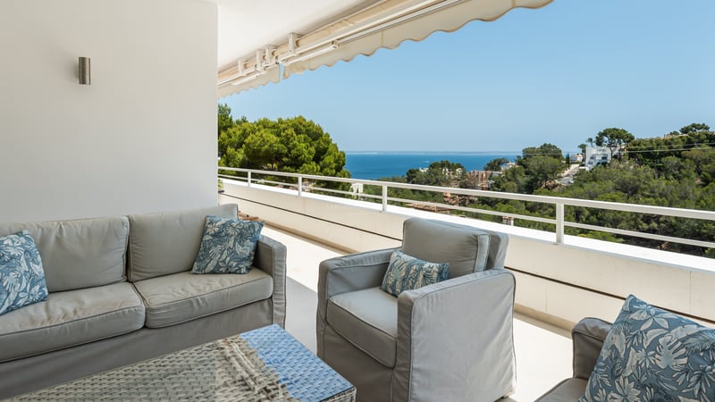 Terrasse couverte avec salon de jardin gris et table en rotin donnant sur la mer et les collines verdoyantes.