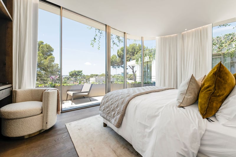 Chambre de luxe avec baies vitrées du sol au plafond, fauteuil en bouclé et vue sur une terrasse privée ensoleillée.