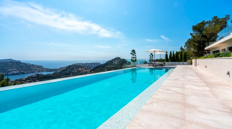 Piscine à débordement turquoise avec vue sur la mer et les montagnes, entourée de dalles en pierre et parasols blancs.