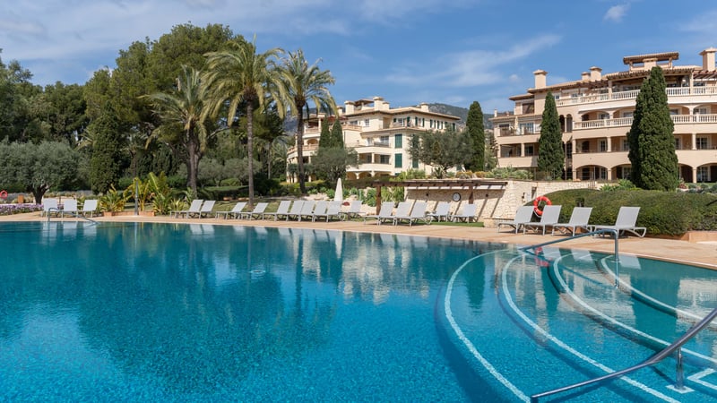 Grande piscine turquoise avec chaises longues et palmiers devant un immeuble d'appartements méditerranéen.