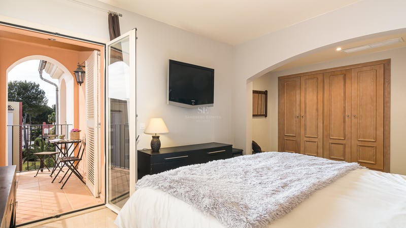 Chambre lumineuse avec placards en bois et portes vitrées ouvrant sur un balcon ensoleillé avec mobilier de bistro.