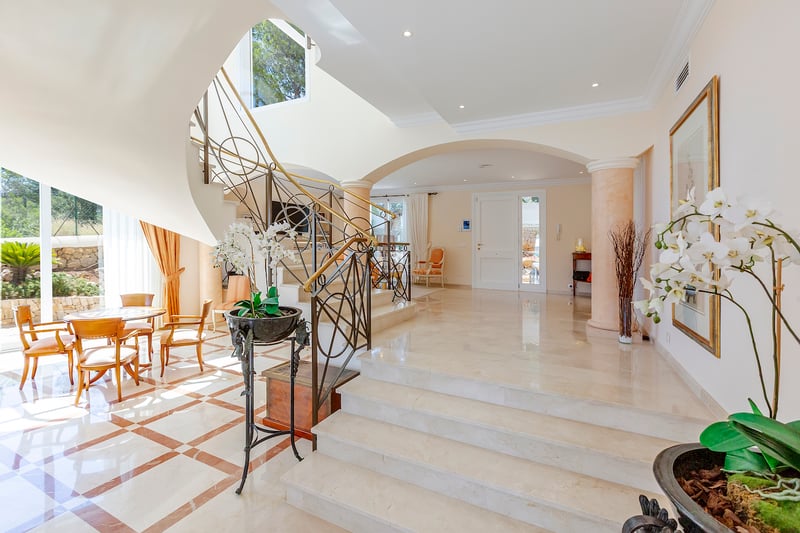 Un hall d'entrée luxueux avec escalier en marbre blanc, rampes en fer forgé et arches architecturales.