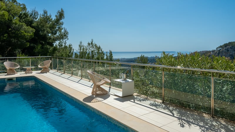 Terrasse de piscine moderne avec garde-corps en verre surplombant les pins et la mer bleue sous un ciel dégagé.