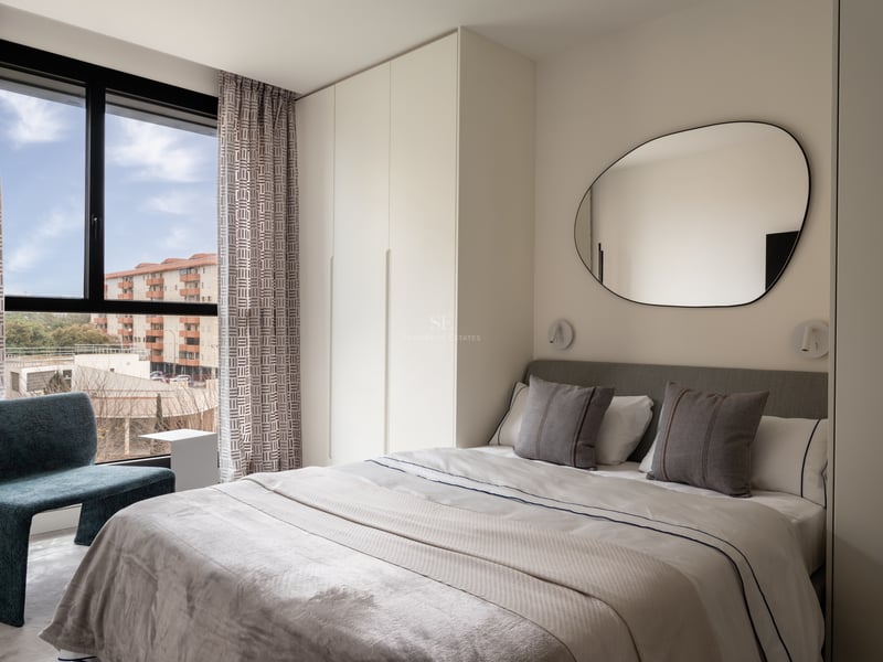 Chambre moderne avec lit blanc, miroir organique, placards intégrés et fauteuil bleu près d'une grande fenêtre.