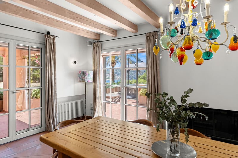 Salle à manger intérieure avec table en bois, lustre coloré, poutres apparentes et portes ouvrant sur une terrasse.