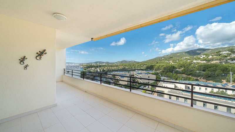 Grand balcon en carrelage blanc avec rambarde noire offrant une vue sur des collines verdoyantes et un ciel bleu.