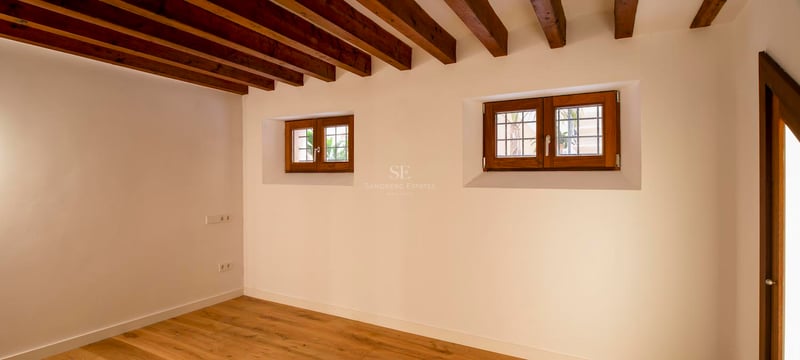 Pièce vide avec plafond à poutres en bois sombre, murs blancs et petites fenêtres en bois.