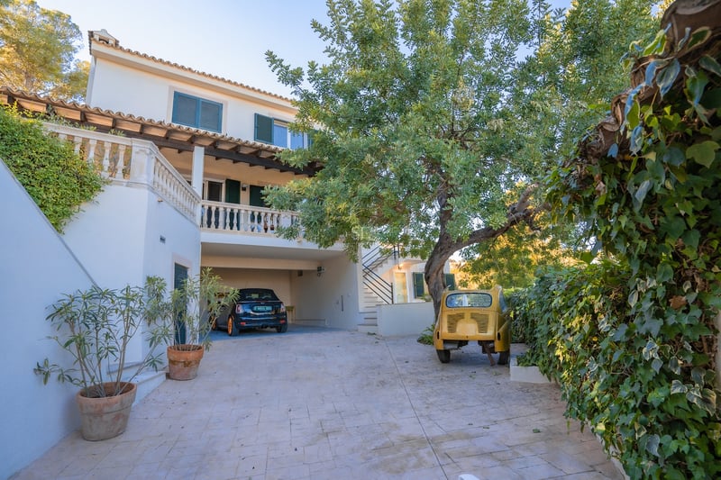 Allée pavée menant à un parking couvert dans une villa blanche avec des volets verts et des arbres.