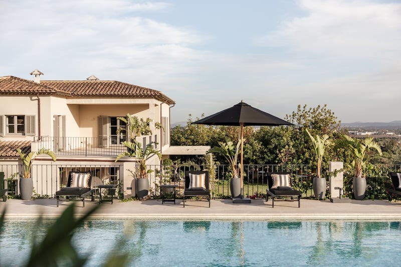 Piscine rectangulaire bordée de bains de soleil noirs, parasol sombre et façade de villa méditerranéenne.