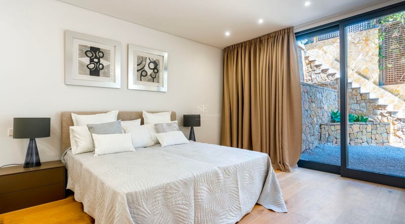 Chambre lumineuse avec lit double, tableaux abstraits et grandes baies vitrées ouvrant sur un patio en pierre.