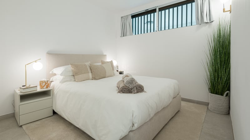 Chambre lumineuse avec lit rembourré beige, linge blanc, lampes en laiton et plante d'intérieur haute.
