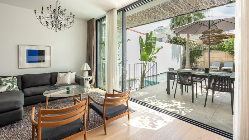 Salon lumineux avec canapé sombre et chaises en bois ouvrant sur une terrasse ensoleillée avec piscine et coin repas.
