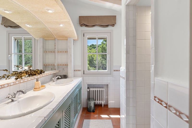 Salle de bain lumineuse avec deux lavabos blancs, meubles verts, sol en terre cuite et vue sur les arbres.