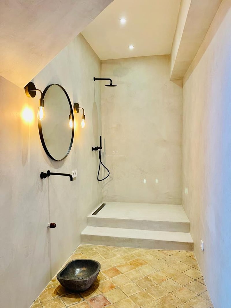 Salle de bain minimaliste avec murs en microciment, sol en terre cuite, vasque en pierre et robinetterie noire mate.