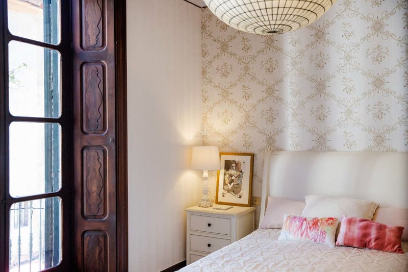 Dormitorio con papel tapiz estampado, cama blanca, lámpara de noche y marco de ventana de madera oscura.
