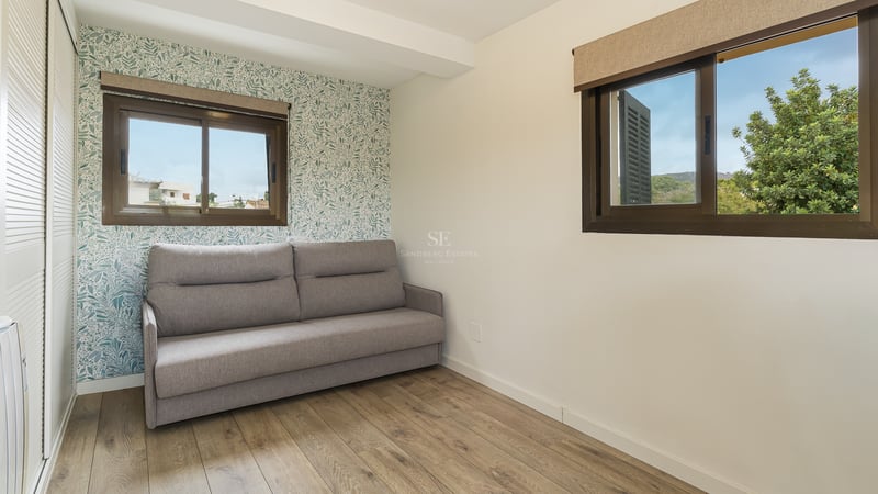 Chambre moderne avec un canapé gris, parquet en bois, papier peint à motif végétal et deux fenêtres.