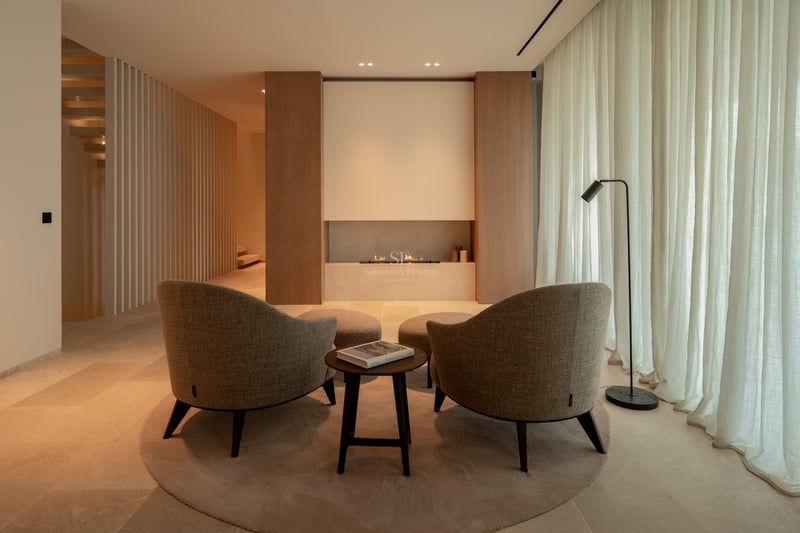 Salon minimaliste avec deux fauteuils beiges face à une cheminée linéaire, éclairage chaleureux et boiseries.