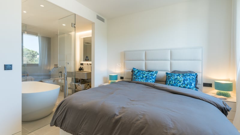 Chambre moderne avec lit gris, lampes turquoise et salle de bain vitrée comprenant une baignoire îlot blanche.
