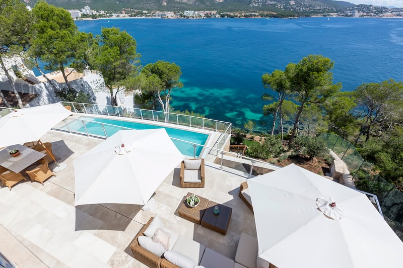 Vue en plongée d'une terrasse en pierre avec parasols blancs, mobilier de salon et piscine surplombant la mer.