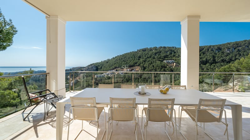 Terrasse couverte moderne avec table blanche et garde-corps en verre, vue sur les collines verdoyantes et la mer.